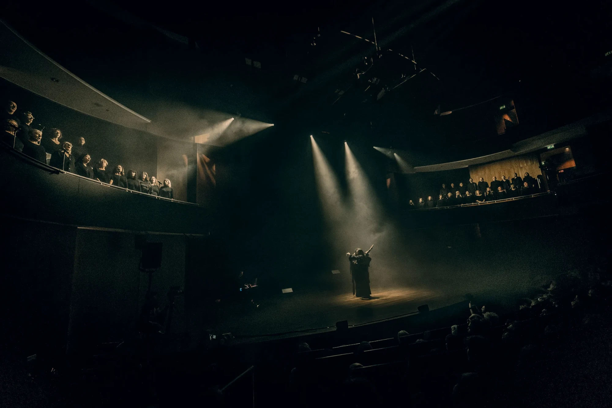 A dancer in a spotlight on the stage of Hertz, with choirs singing from the balconies on both sides